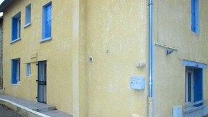 Een geel gebouw met blauwe raamkozijnen, een kleine deur en een meterkast aan de zijkant.