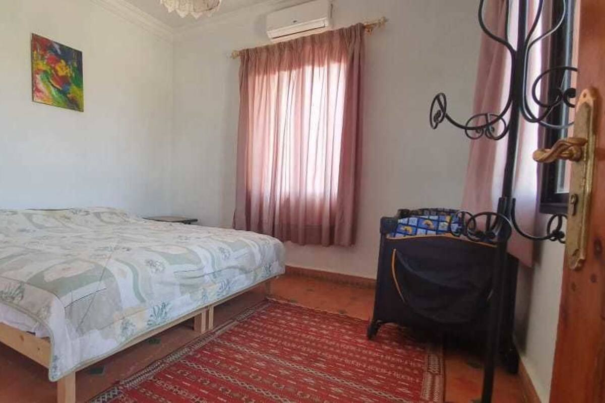 Un dormitorio con cama, una mesita, una silla, una alfombra roja, una ventana con cortinas y una puerta con pomo metálico.