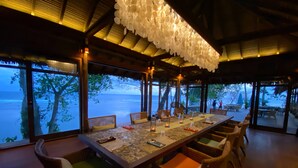 A dining area with a long table, wicker chairs, and a view of the sea.
