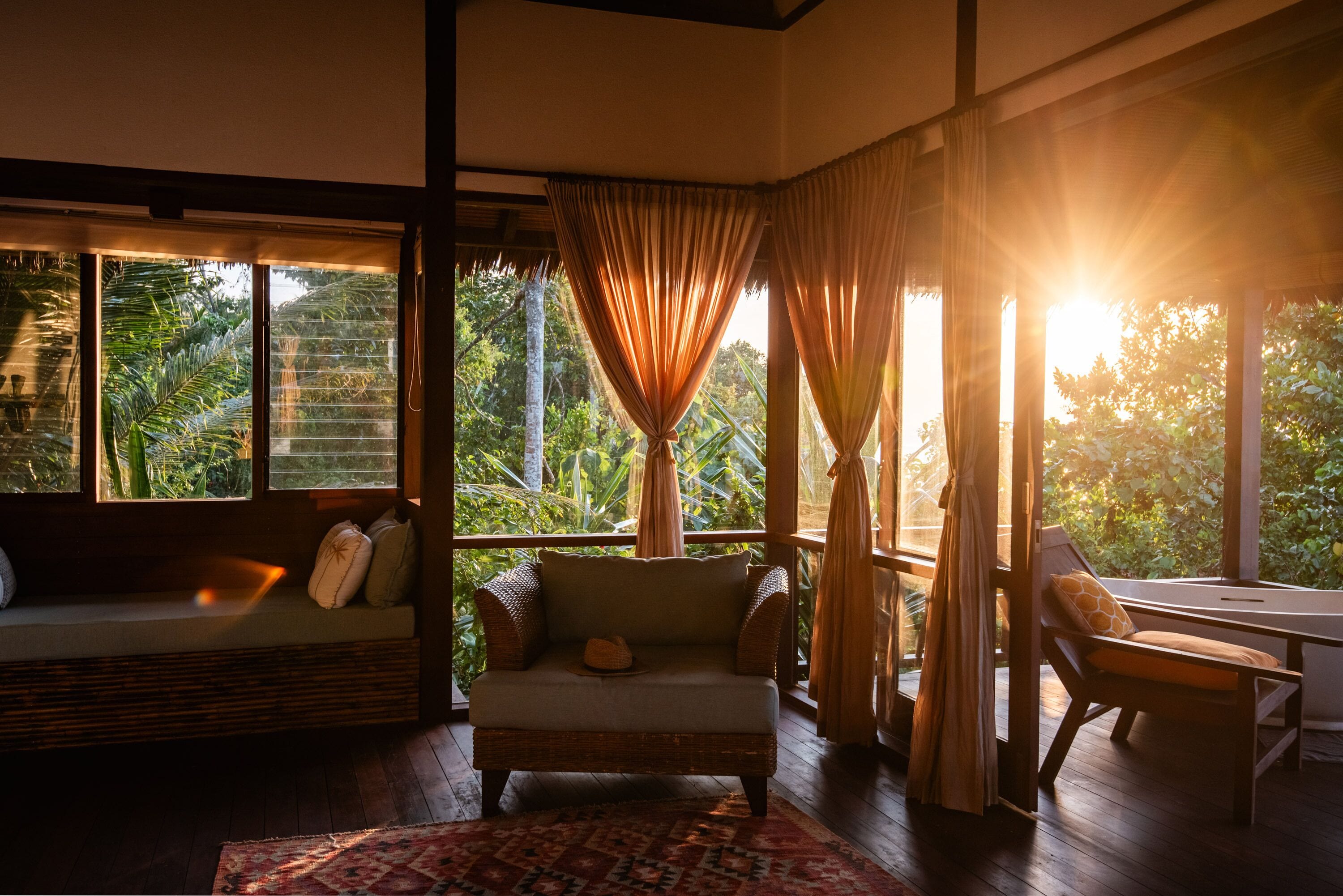 A room with a sofa, a chair, and a table. There are windows with curtains, and a view of greenery outside.