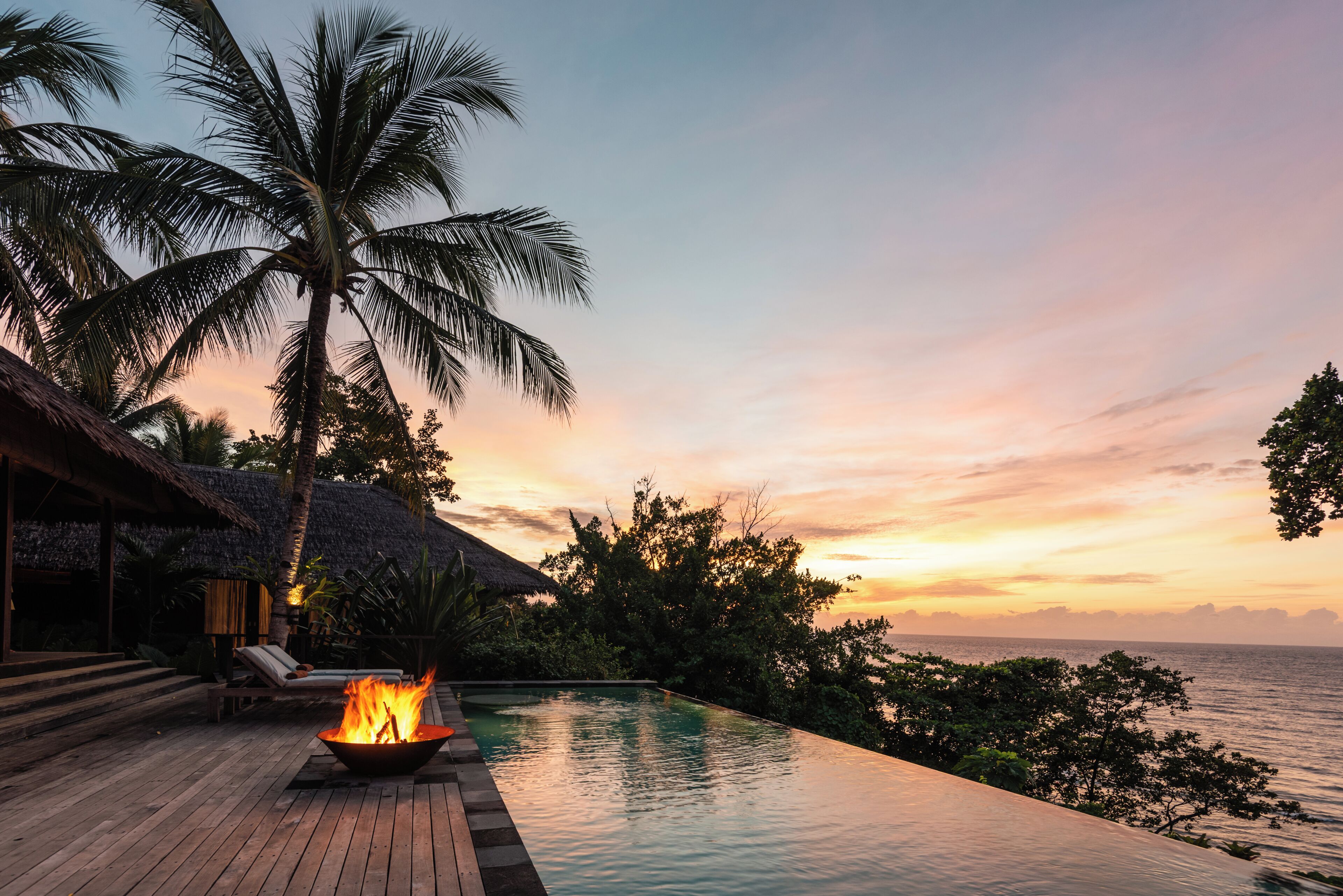 A wooden deck with a fire feature, overlooking a pool and the sea at sunset.