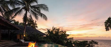 A wooden deck with a fire feature, overlooking a pool and the sea at sunset.