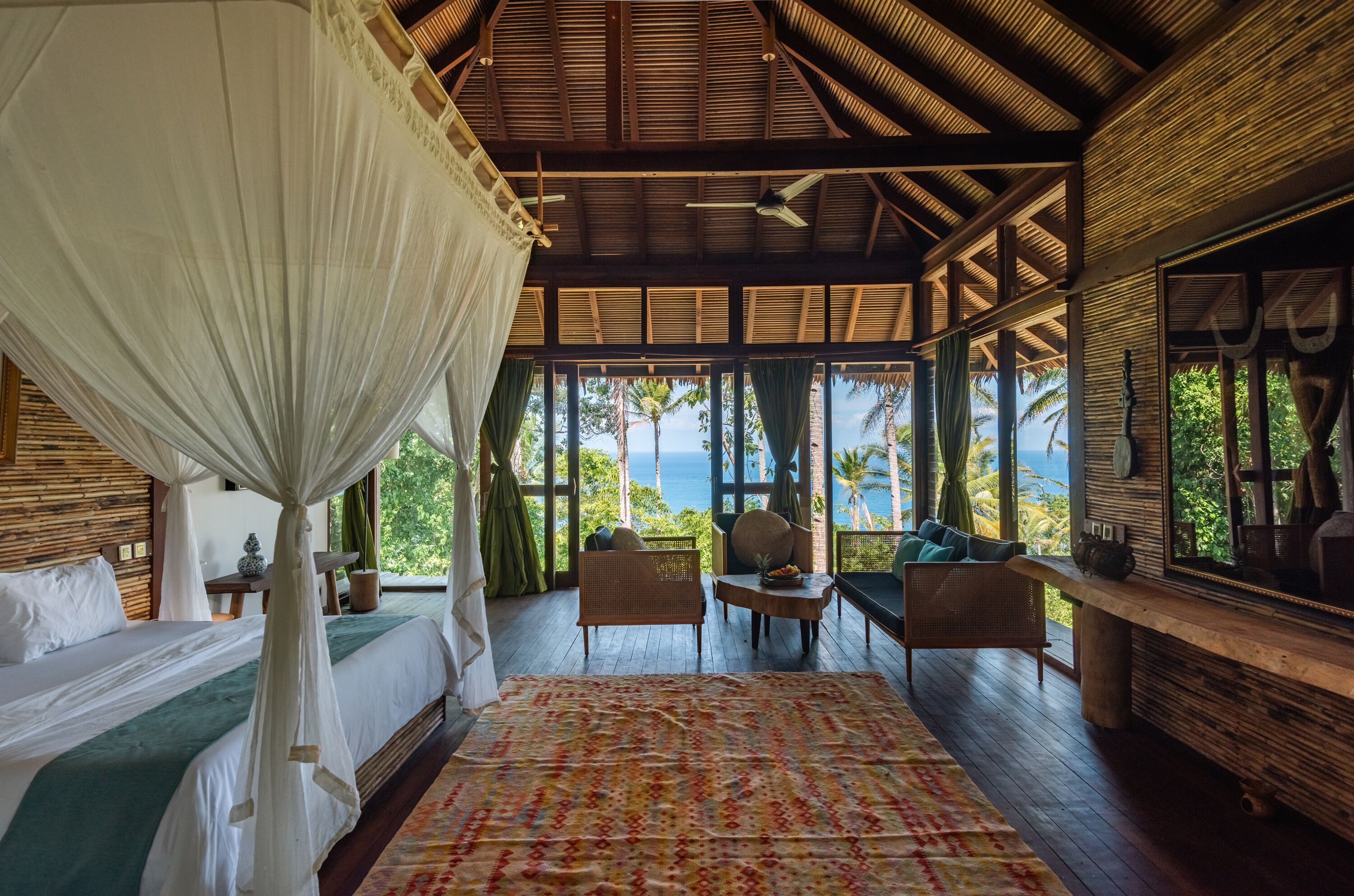 A room with a bed, a table, and chairs, a large window with a view of the ocean, and a ceiling fan.