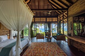 A room with a bed, a table, and chairs, a large window with a view of the ocean, and a ceiling fan.