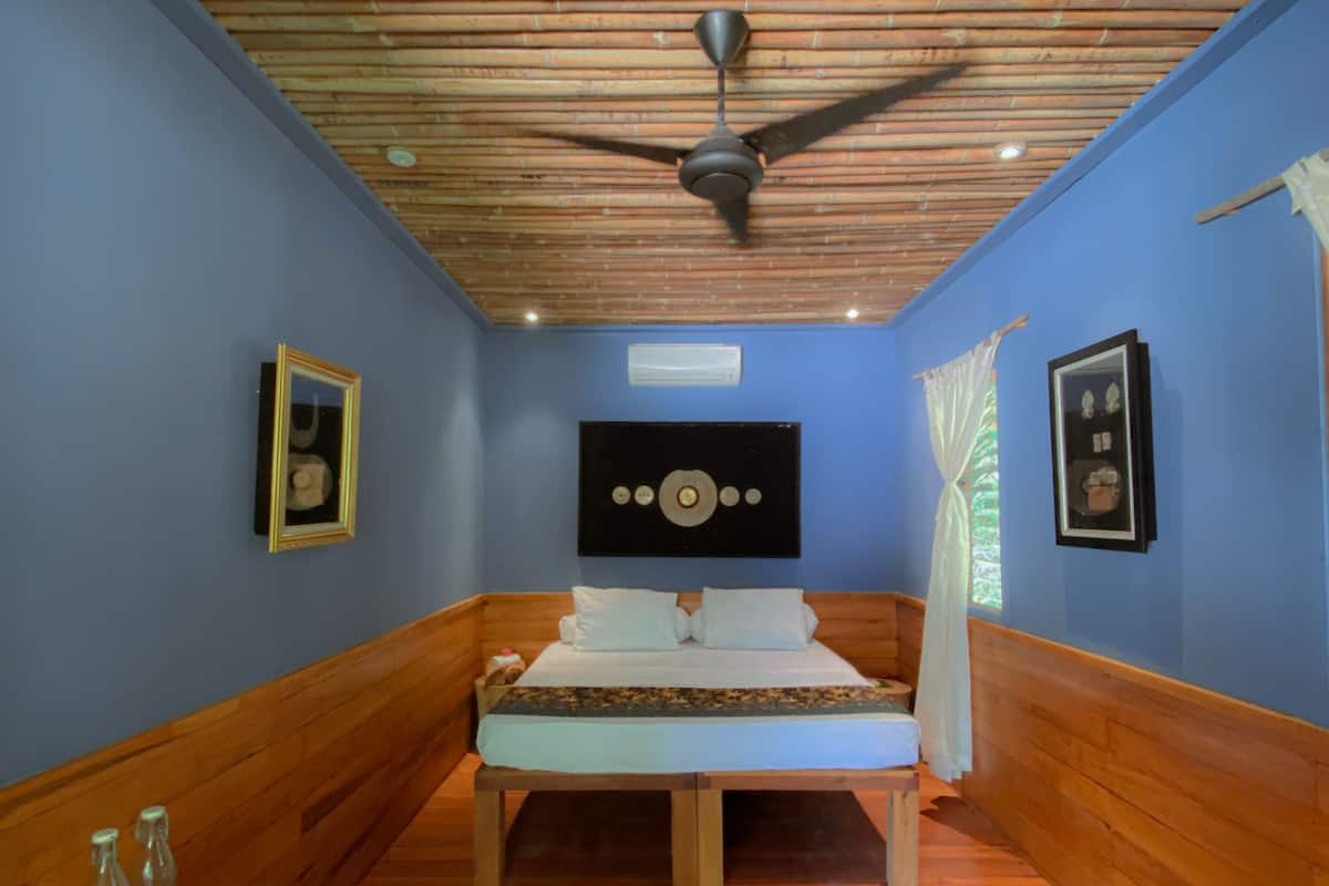 Um quarto pequeno com uma cama, paredes de madeira, um ventilador de teto e quadros nas paredes.