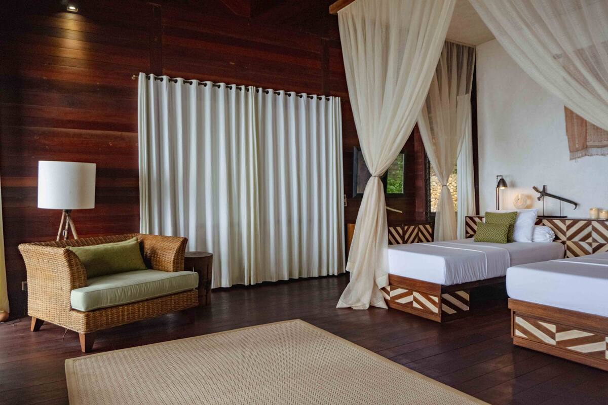 Um quarto com piso de madeira, um sofá de vime, duas camas com cabeceiras estampadas, uma janela grande com cortinas e um abajur.