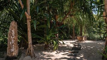 Tropische tuin met een houten bord waarop 'Hill Top Cabin' staat, omringd door weelderige begroeiing en een zandpad dat naar een zitgedeelte leidt.