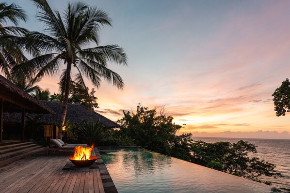 A wooden deck with a pool, a fire pit, and a thatched-roof building.