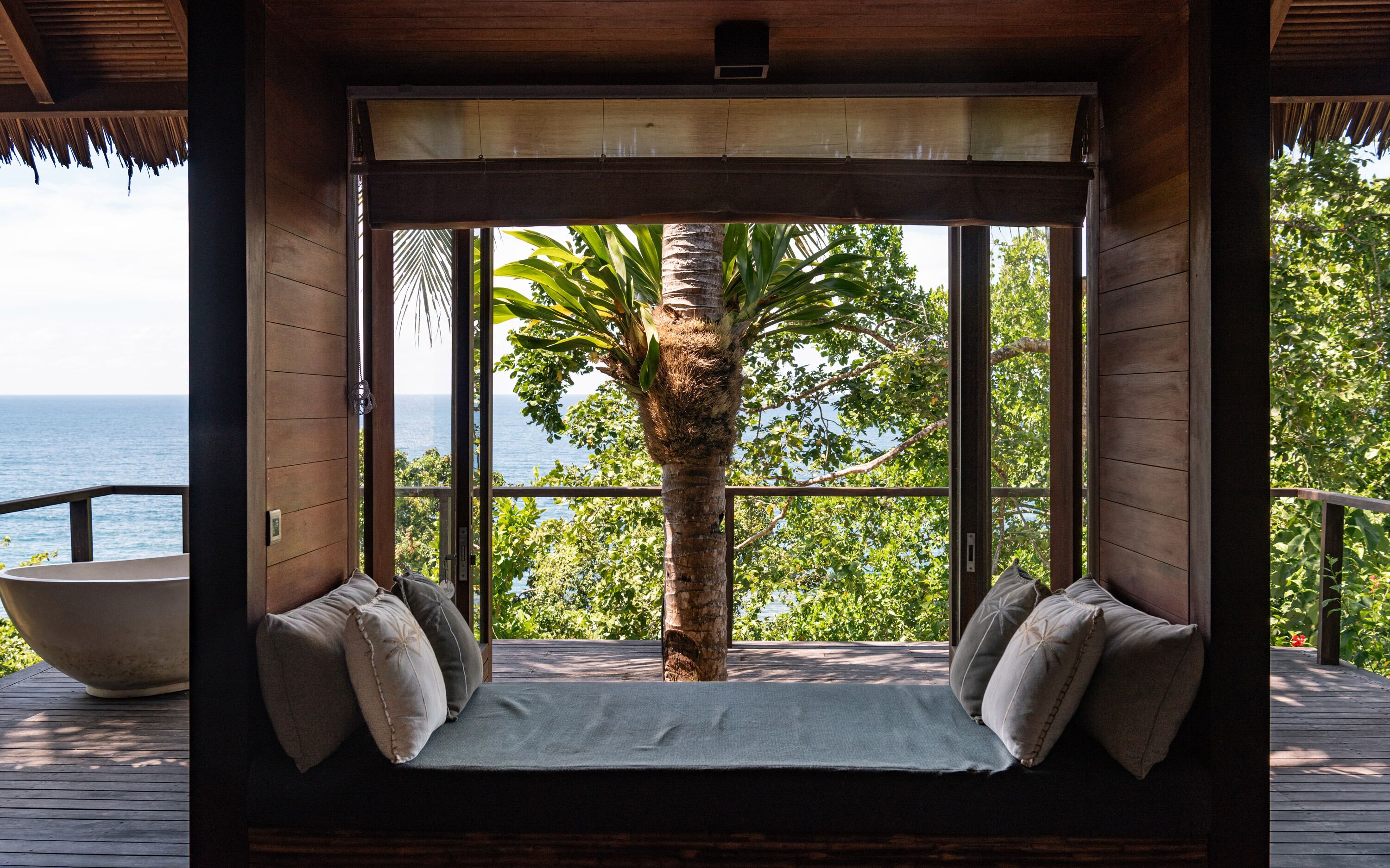 A wooden deck with a built-in seating area featuring a cushioned bench and pillows, overlooking a lush green landscape and the sea.
