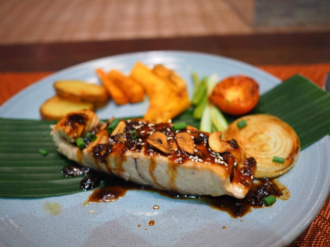 Ein Teller mit gegrilltem Fisch, bedeckt mit Sauce und garniert mit Frühlingszwiebeln und Sesamsamen, dazu frittierte Beilagen und eine ganze Zwiebel.