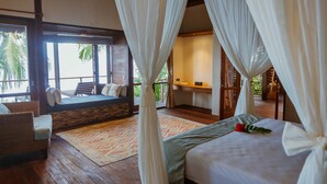 A bedroom with a bed, a sitting area with a chair and a sofa, a wooden desk, and a large window with curtains.