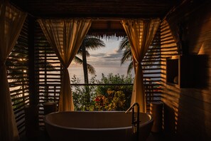 Een badkamer met een bad, gordijnen en uitzicht op zee en palmbomen.