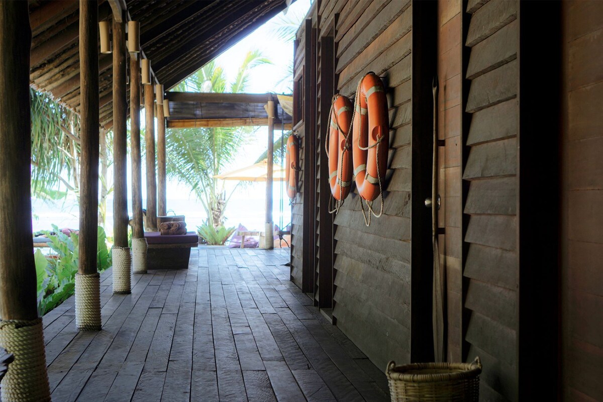 A covered wooden walkway with a tiled floor, a woven basket, and a bench with cushions.