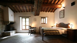 A rustic bedroom with exposed wooden beams, a stone fireplace, a wooden bed, a desk, and two windows.