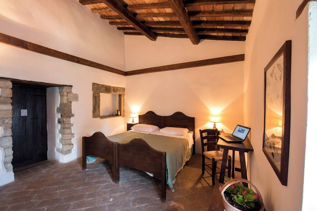 A bedroom with a wooden bed, a desk with a laptop, a stone wall, and a rustic ceiling.