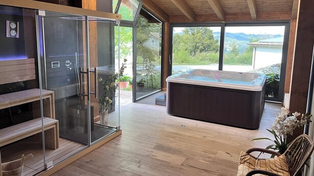 A modern bathroom with a glass shower enclosure, a wooden bench, a hot tub, and a view of the outdoors.
