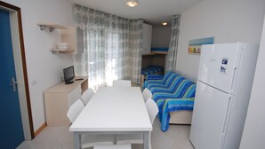 A compact living space with a white table and chairs, a kitchenette with a refrigerator, and a bed with a striped blanket.