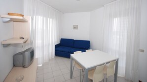 A small, bright room with a white table, chairs, a blue sofa, a television, and a ceiling light.