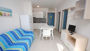 A compact room with a bed, a small dining table with chairs, a kitchenette, and a TV on a desk.