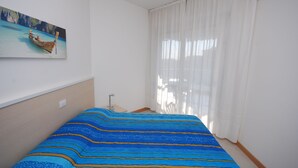 A bedroom with a bed, a painting of a boat on the wall, and a window with curtains.