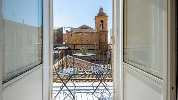 A balcony with a view of a building with a bell tower, metal chairs, and a small table. - B&B Home Visit (Agrigento)