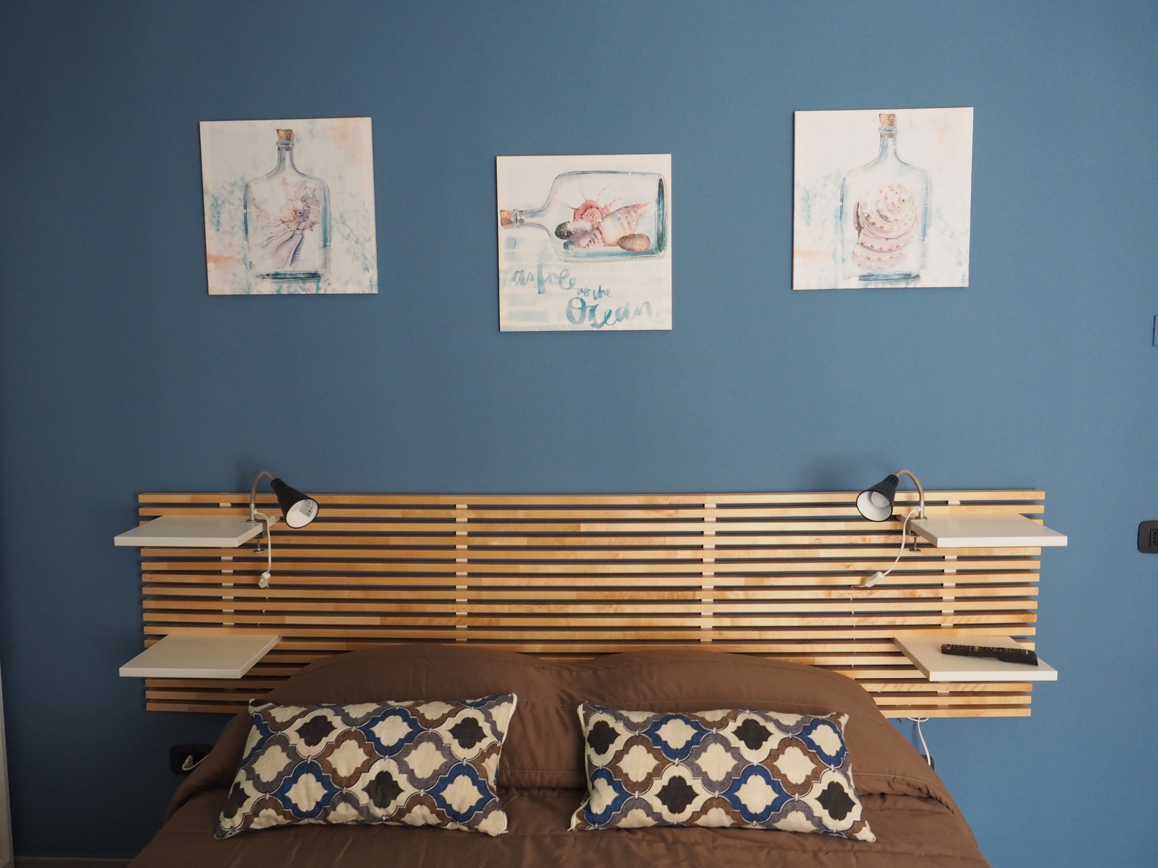 Ein Schlafzimmer mit einem Lattenrost-Kopfende, zwei Kissen, einem Nachttisch und drei an der Wand befestigten Bildern mit Darstellungen von Flaschen.