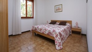 A bedroom with a wooden bed, white walls, a window with curtains, a bedside table with a lamp, and a framed picture on the wall. - Residence Orsa Maggiore (Lignano Sabbiadoro)