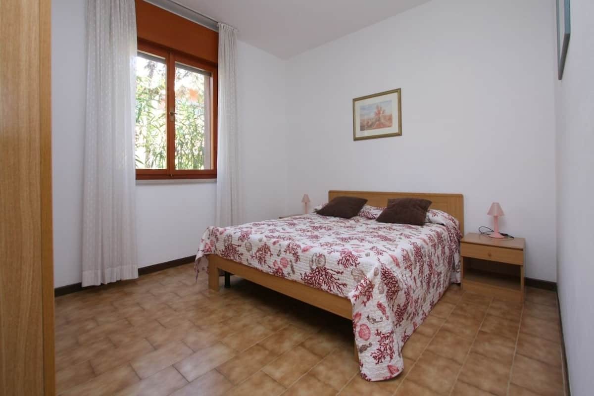 A bedroom with a wooden bed, white walls, a window with curtains, a bedside table with a lamp, and a framed picture on the wall.