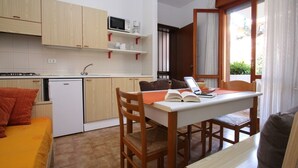 A compact kitchen with wooden cabinets, a microwave, and a small refrigerator. There's a dining area with a table and chairs, and a bed with an orange bedspread. - Residence Orsa Maggiore (Lignano Sabbiadoro)