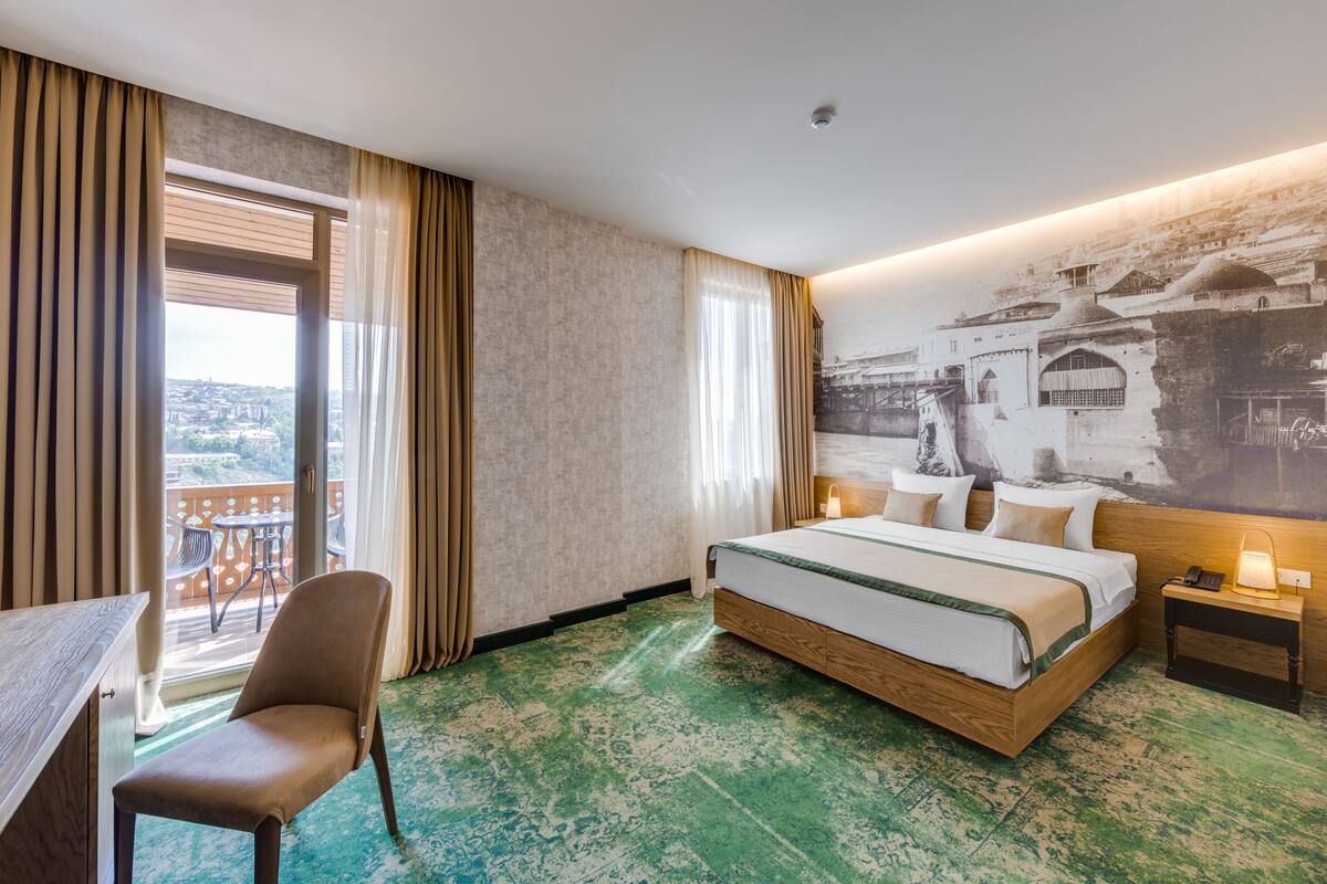 A hotel room with a large bed, a desk, a chair, and a balcony with a view of the city.