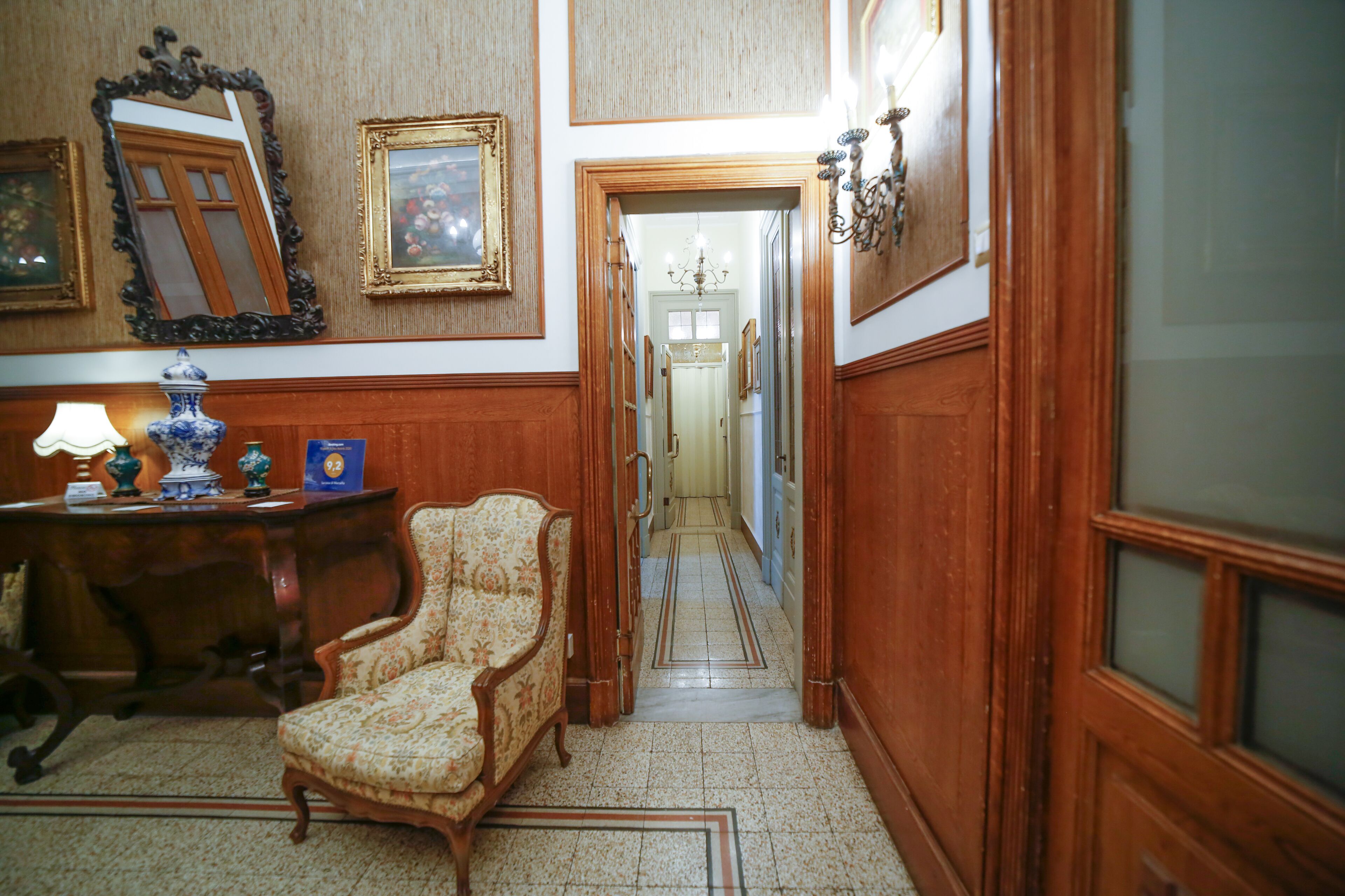 A hallway with a wooden floor, a chair, a desk with a lamp, and framed pictures on the walls.