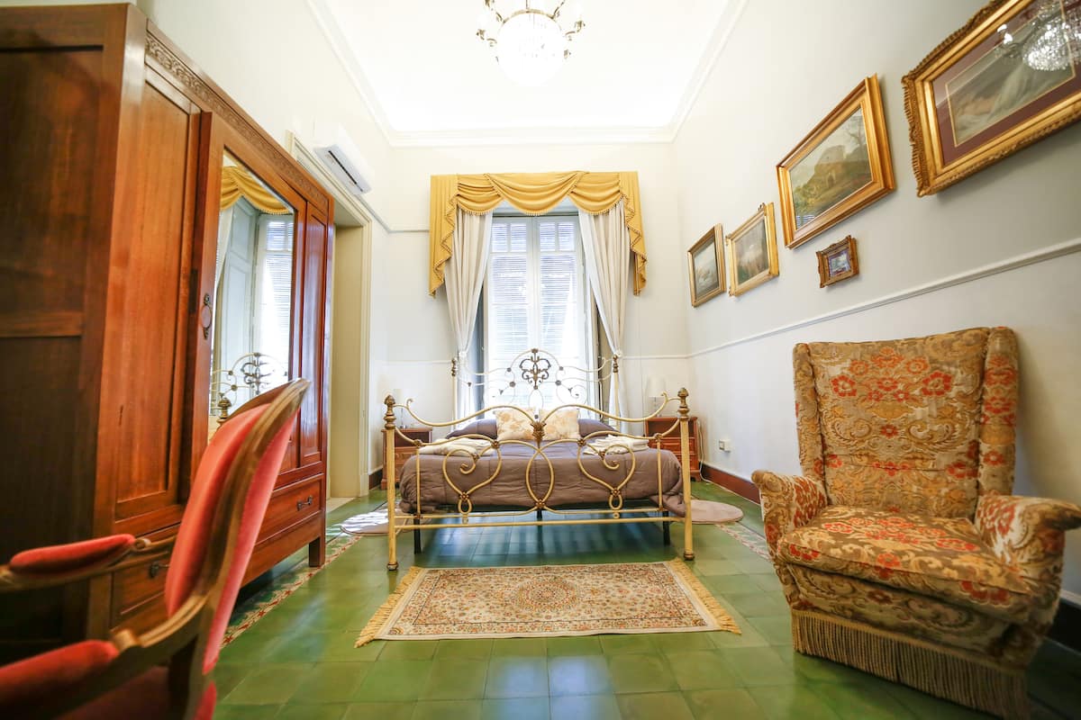 A bedroom with a four-poster bed, a wooden wardrobe, a floral armchair, and a patterned rug.