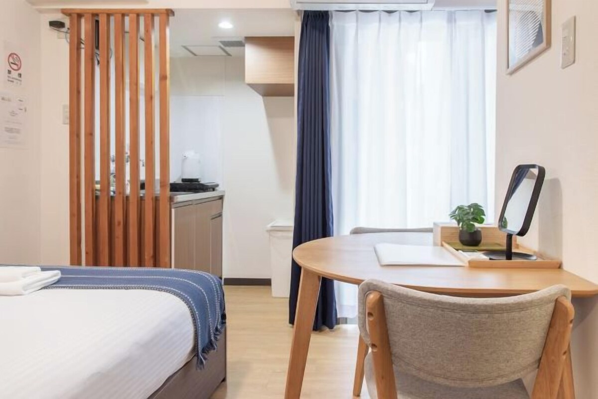A hotel room with a bed, a desk, a chair, a window with curtains, and a view of the kitchen area.