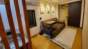 A modern living room with a brown sofa, a small table, and a white rug. There is a window with a black frame, and an air conditioning unit mounted on the wall. - Sakuranomiya Hotel-Self Service (Osaka)