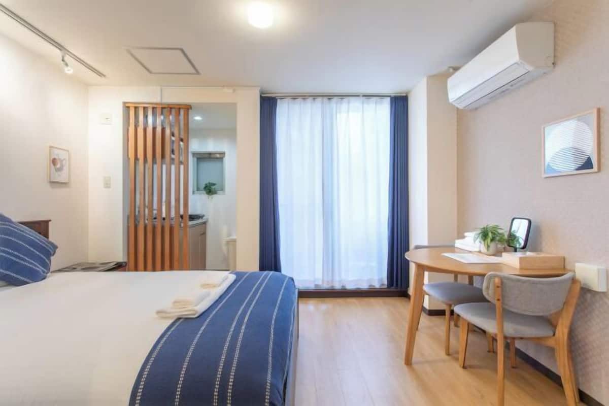 Quarto de hotel com cama, escrivaninha, cadeira, janela com cortinas e ar condicionado.