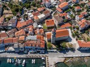 An aerial view of a coastal town with terracotta-roofed buildings, a marina with boats, and a prominent church.