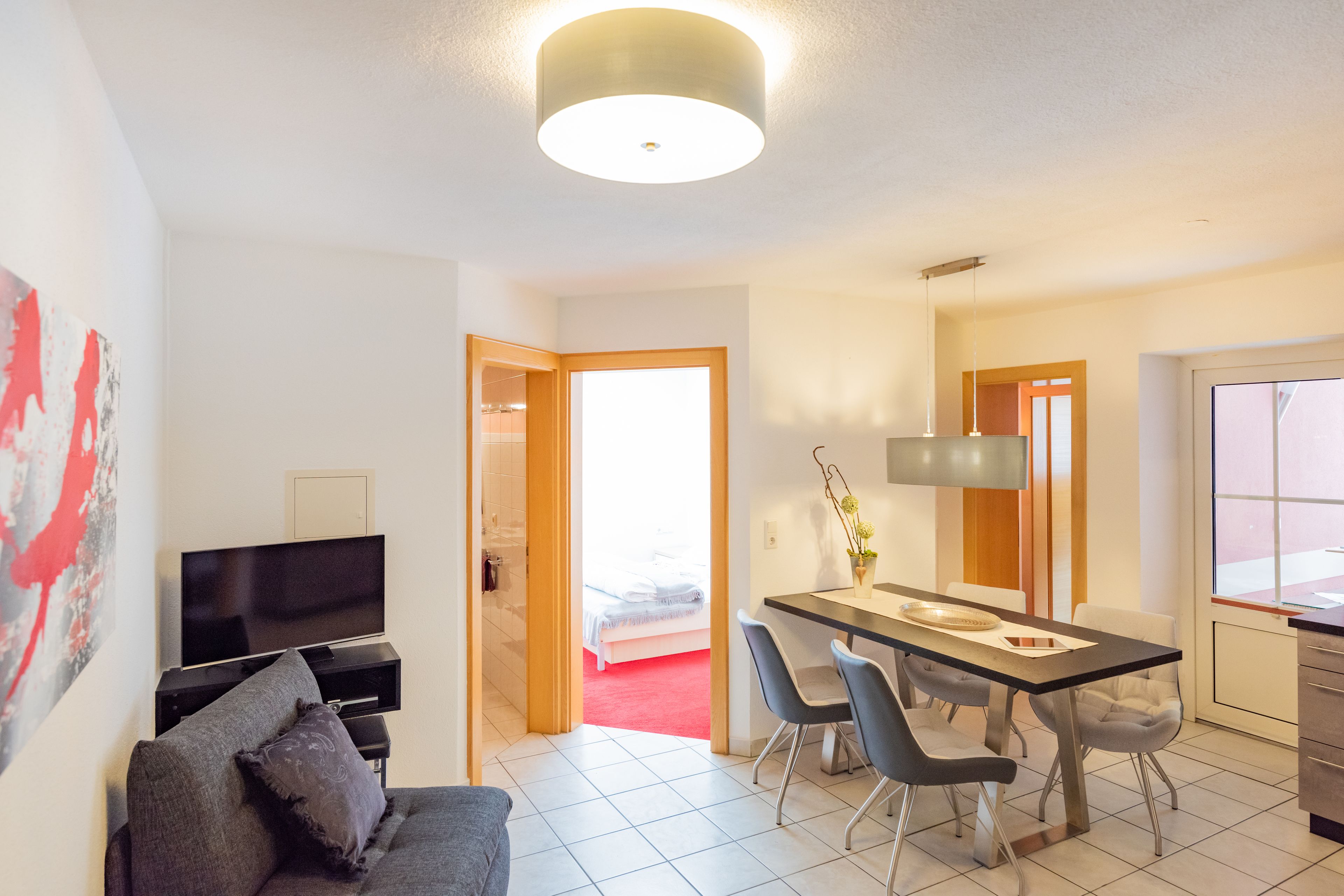 A modern hotel room with a flat-screen TV, a sofa, a dining table with chairs, and a large window.