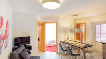A modern hotel room with a flat-screen TV, a sofa, a dining table with chairs, and a large window.