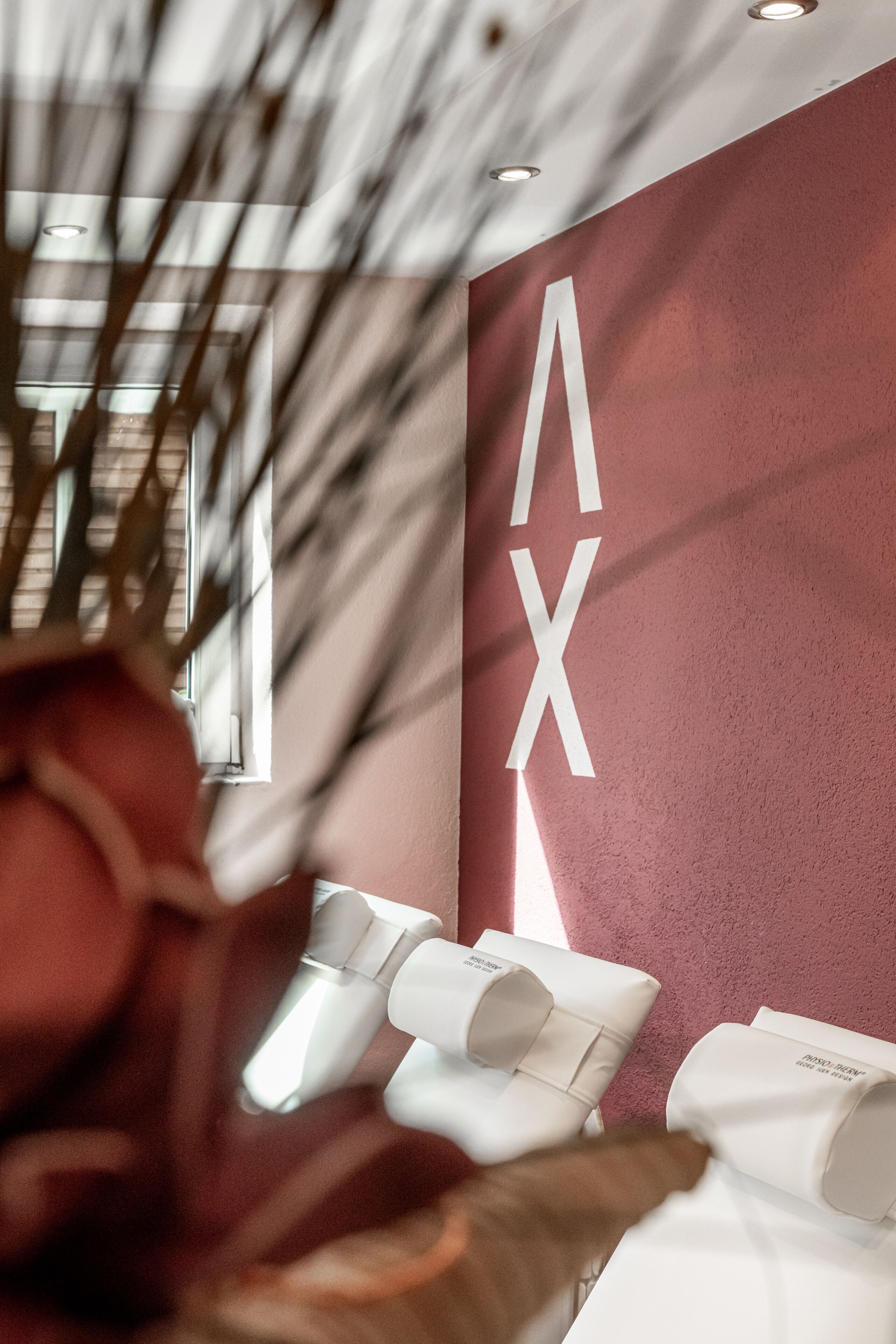 A modern interior with a red wall and a large white 'AX' sign. There are white chairs with a logo on them, and a window allowing natural light to enter.