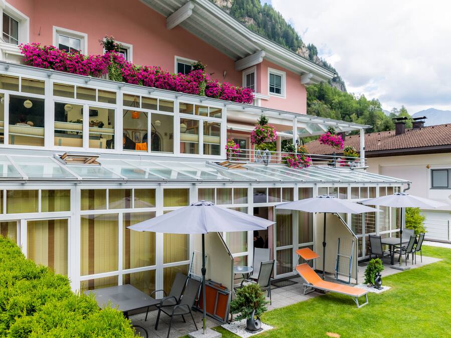 Un bâtiment à deux étages doté d’une véranda en verre, d’un espace extérieur aménagé avec des parasols et d’un jardin bien entretenu.