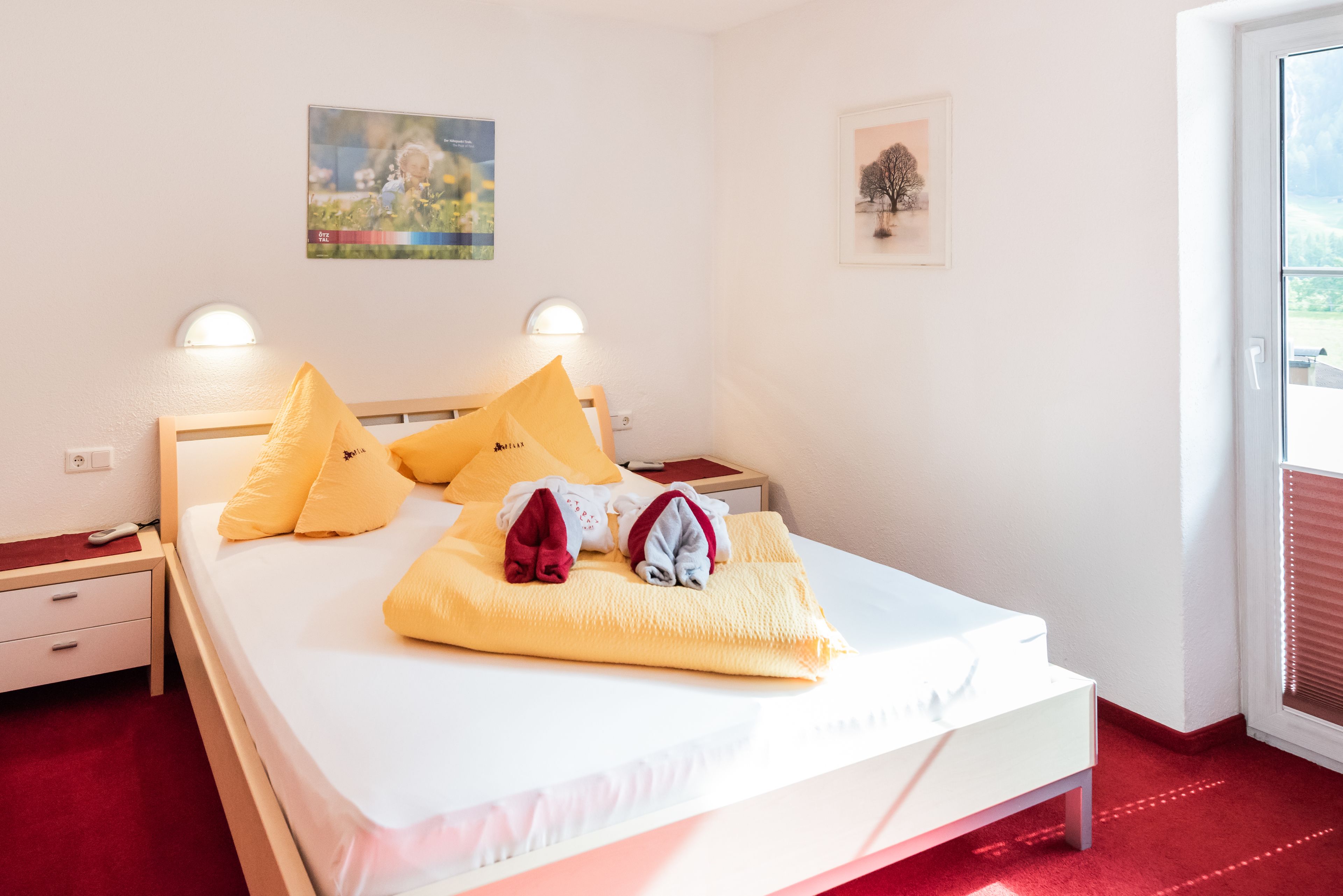 A neatly made bed with yellow pillows and a blanket, a bedside table with a lamp, and a window with blinds.