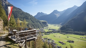 A mountain lodge with a wooden deck and a flag, overlooking a valley with houses and a clear sky. - Apart Relax (Laengenfeld)