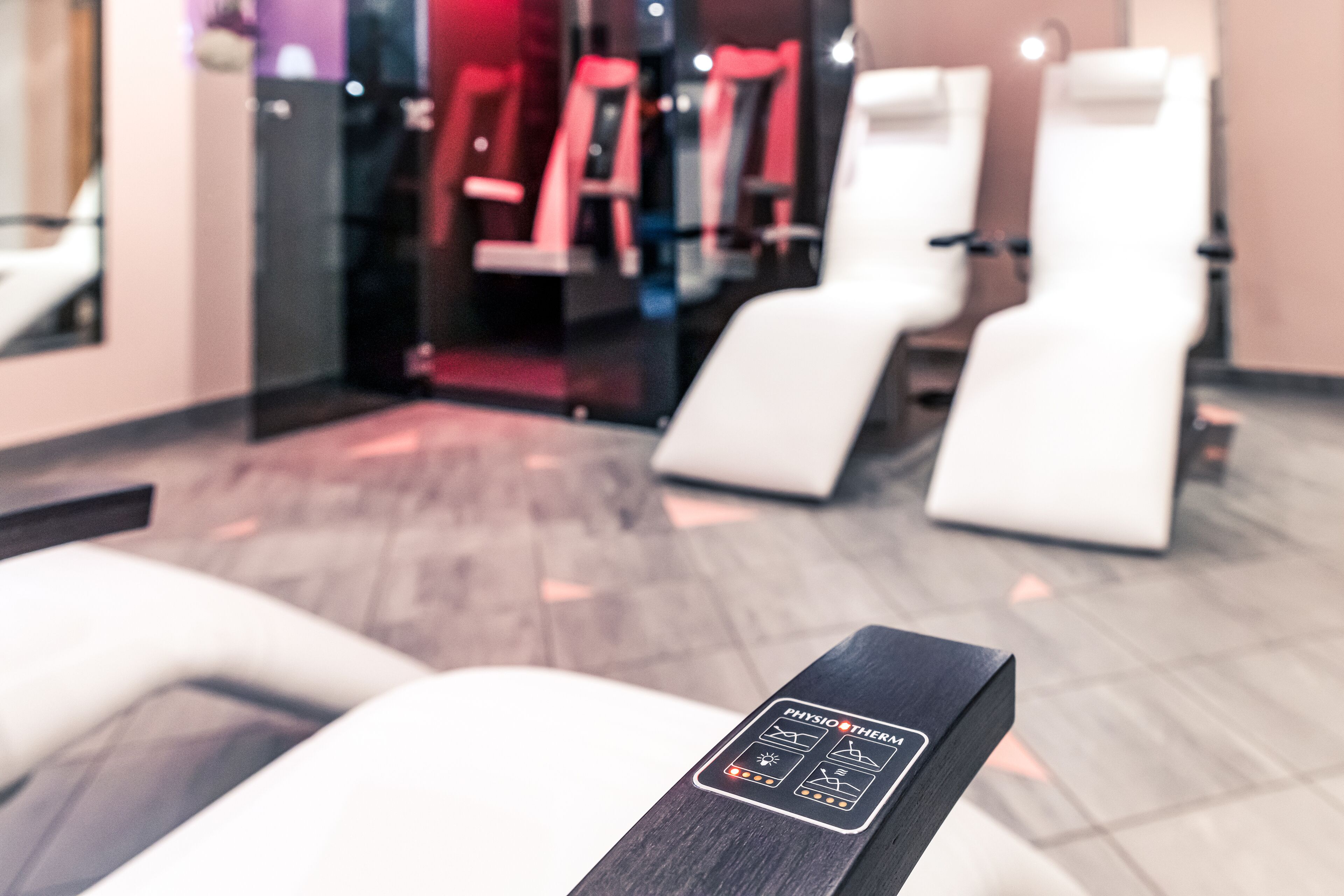 A modern spa room with a massage chair, a wall-mounted massage table, and exercise equipment in the background.