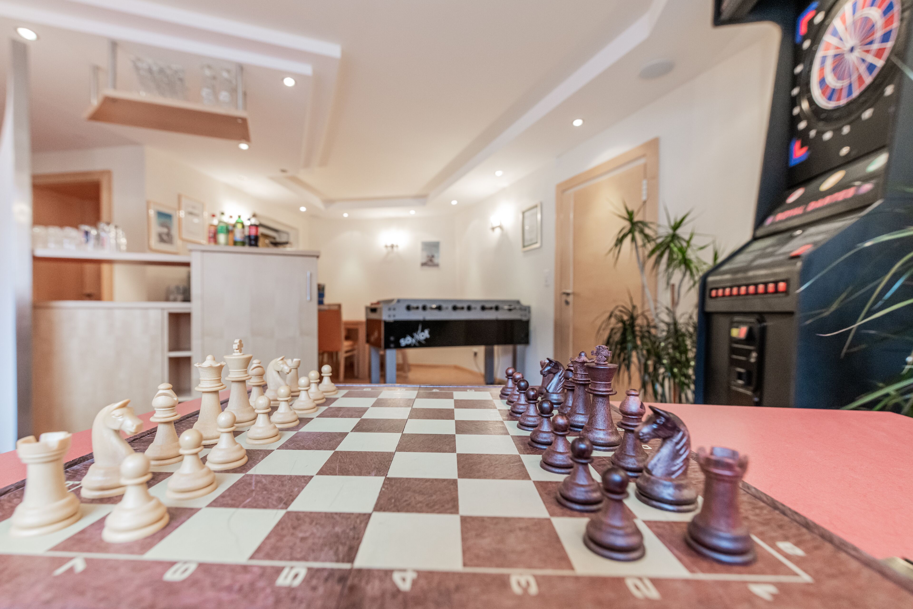 A checkered chessboard with pieces set up in the middle of a room with a dartboard and a jukebox in the background.