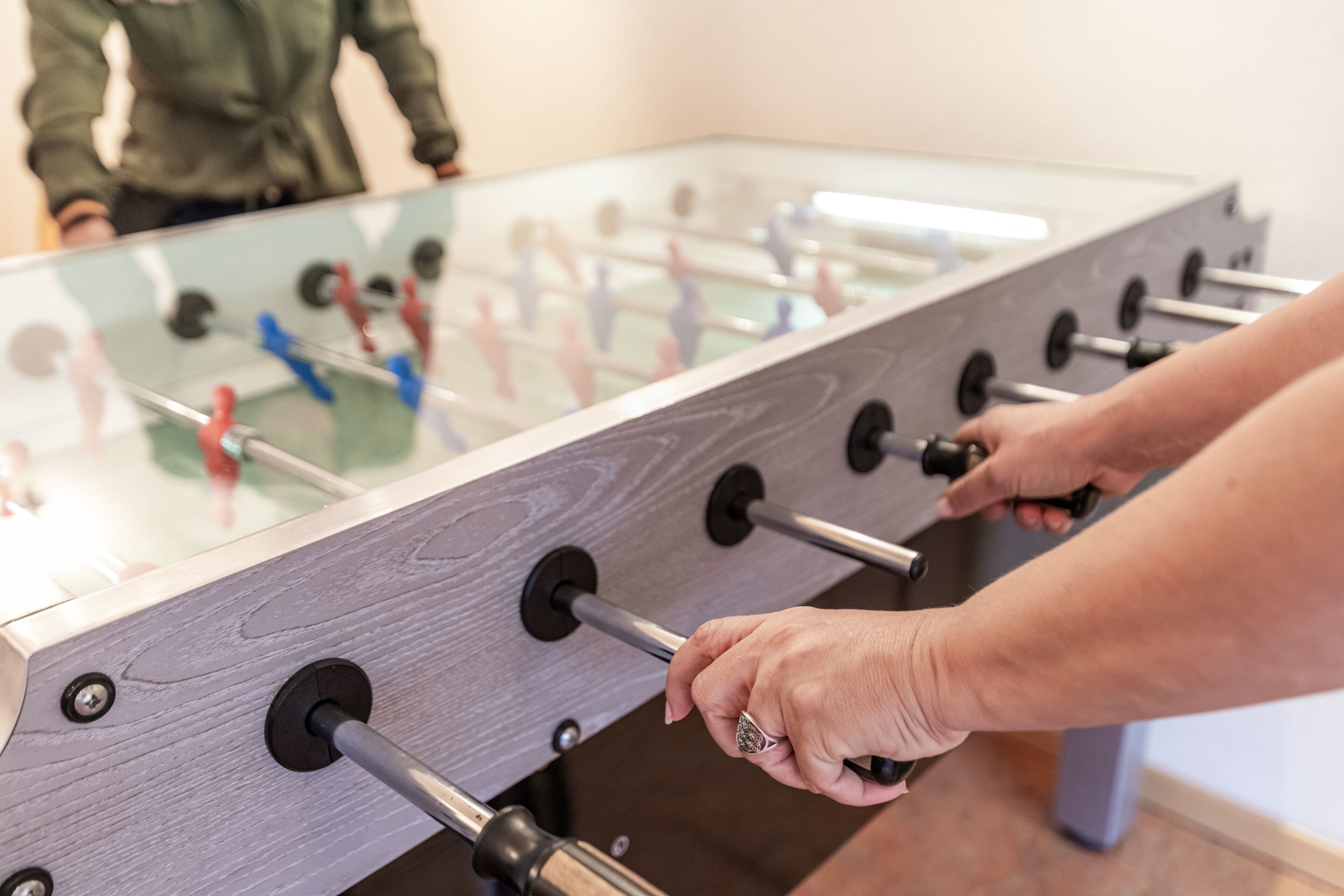 A foosball table with a glass top and metal rods, a person adjusting the rods, and a blurred figure in the background.