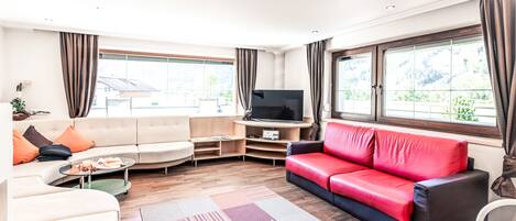 Una sala moderna con un sofá color beige, un sofá de cuero rojo, un televisor de pantalla plana y ventanas amplias con cortinas.