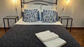 A neatly made bed with a grey blanket, two pillows with a blue and white pattern, and two bedside tables with wall-mounted lamps.