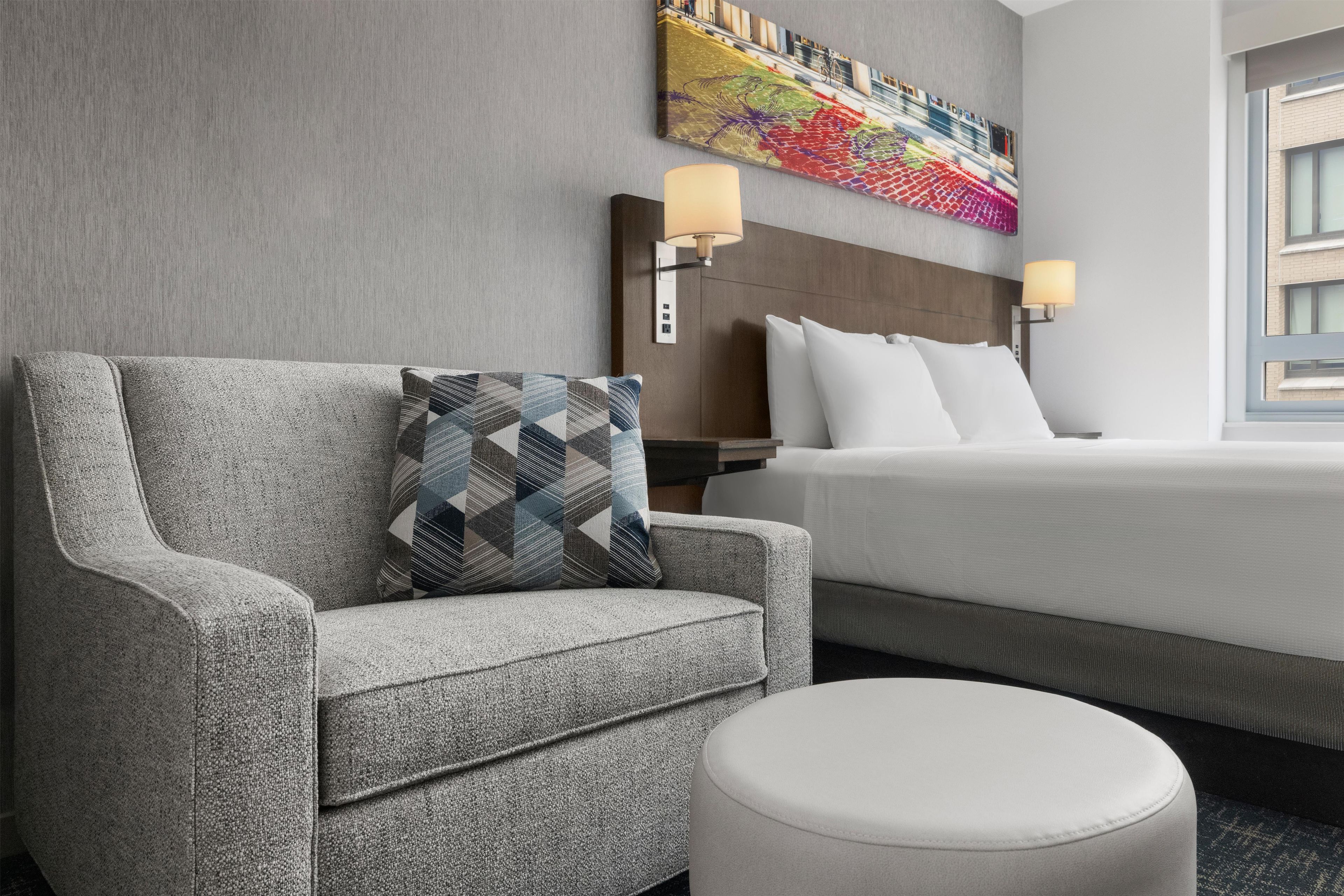 A hotel room with a grey armchair, a patterned pillow, a white ottoman, a bed with white linens, and a colorful abstract painting on the wall.