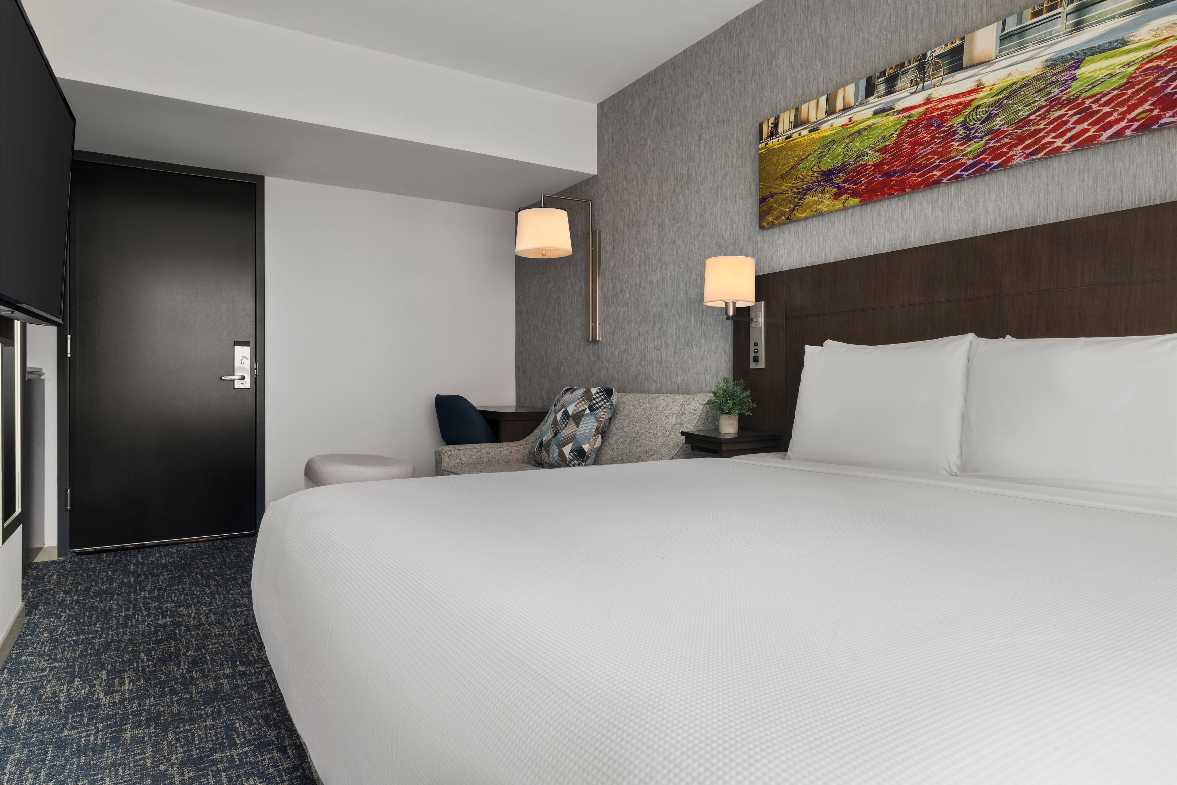 A hotel room with a large bed, a desk, a chair, a lamp, and a painting on the wall.