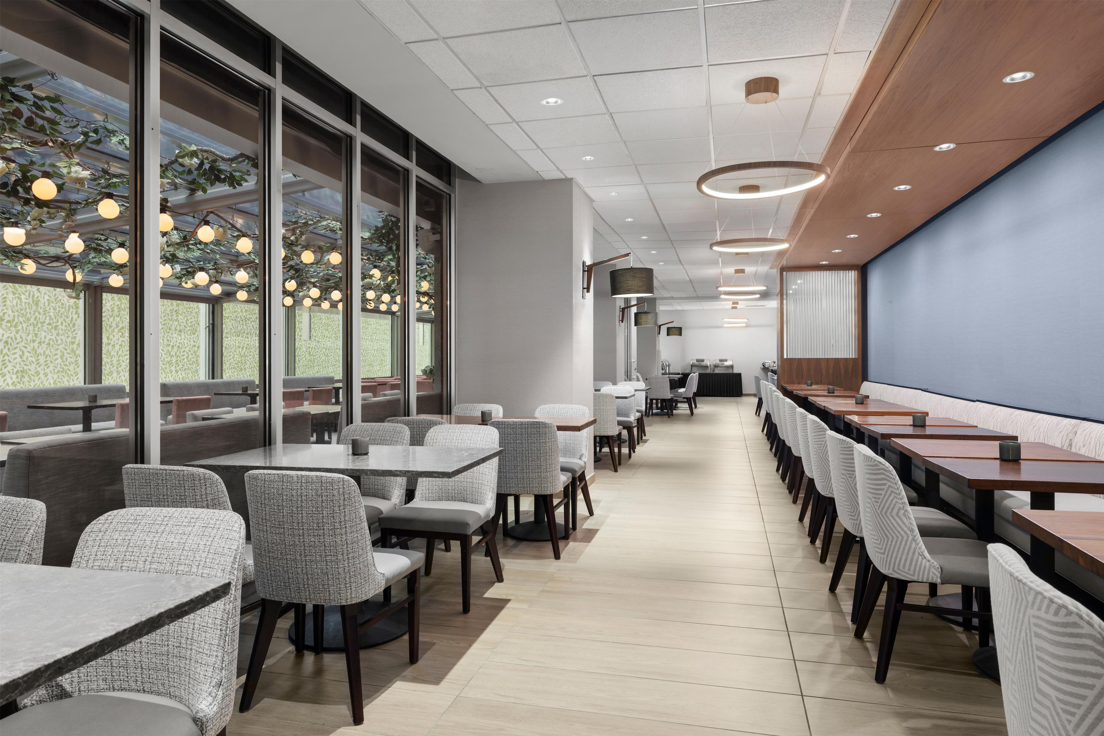 A modern restaurant with long tables and chairs, large windows, and a ceiling with recessed lighting.